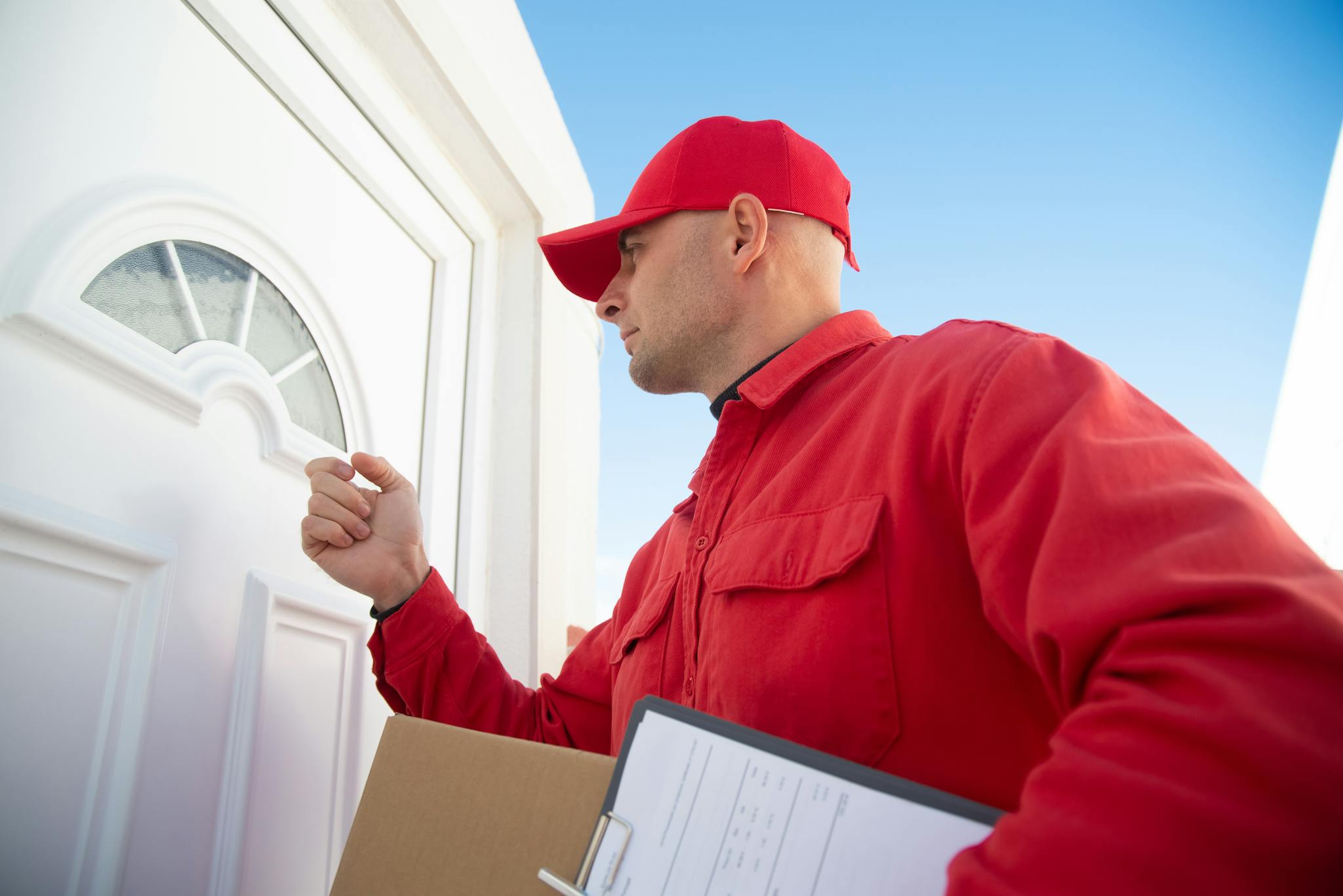 A delivery man in red uniform knocking on a white door carrying a package.
