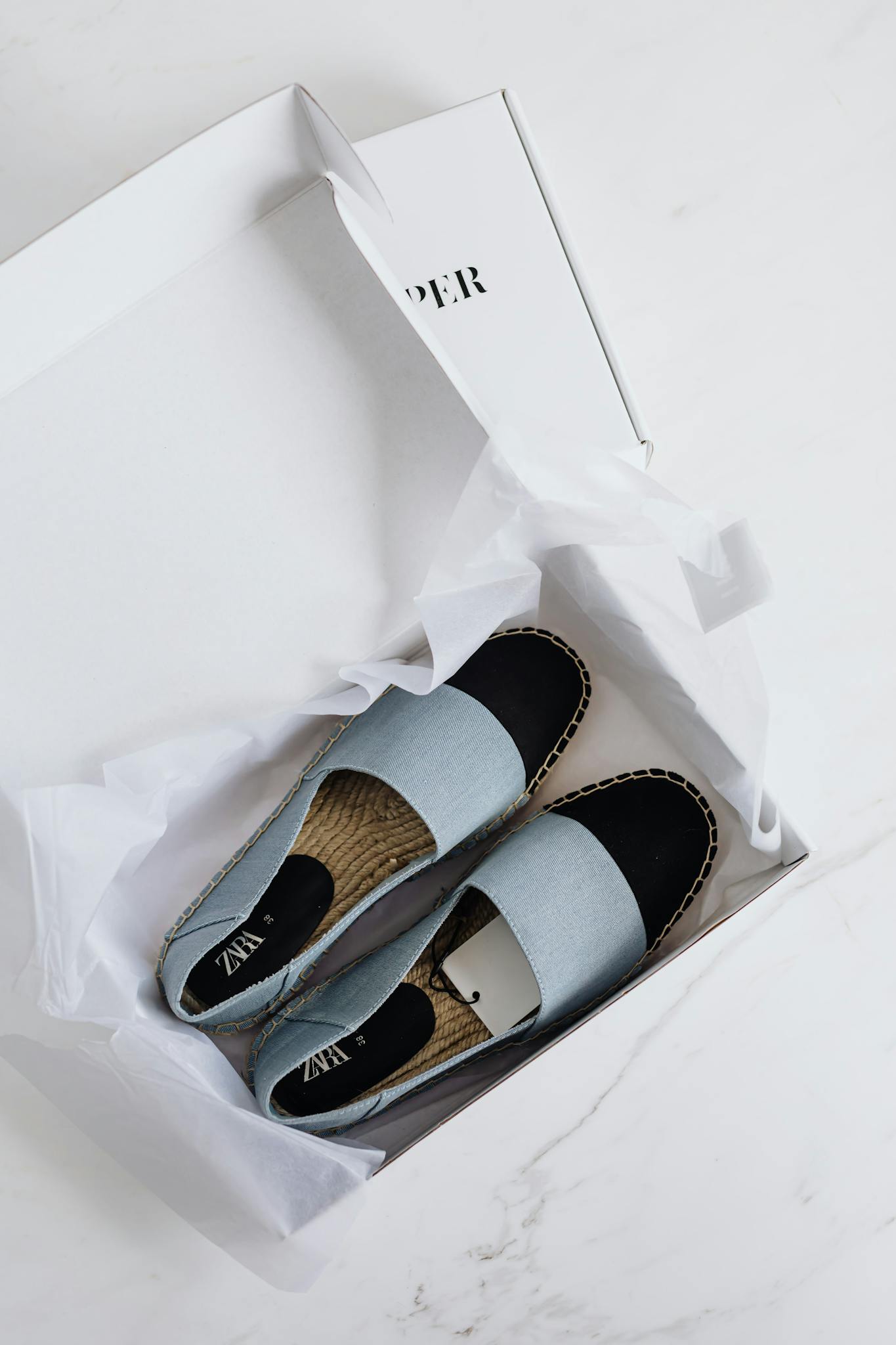 Top view of fashionable espadrilles in an open white cardboard box on a marble surface.
